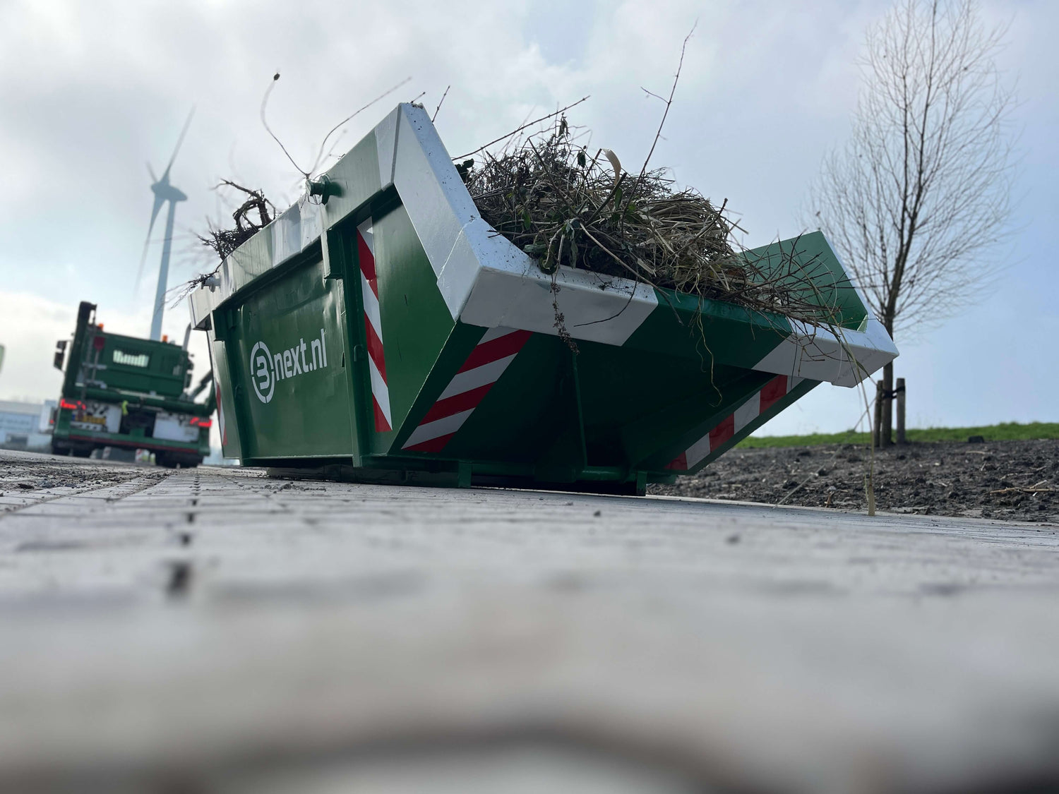 Afvalcontainer gevuld met tuinafval en portaalwagen erbij